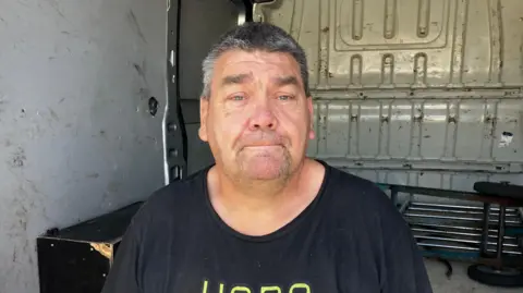 A man with dark, greying hair looks as the camera as he stands against the inside of a van.