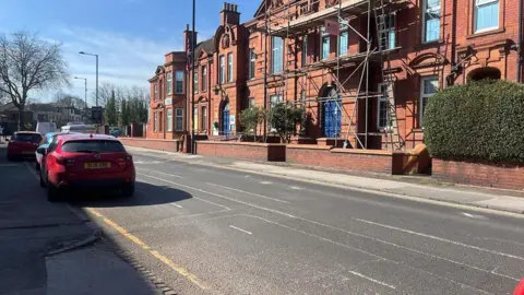 BBC A residential road with a red car parked on the left side and red brick buildings on the right. The sky is blue in the background.