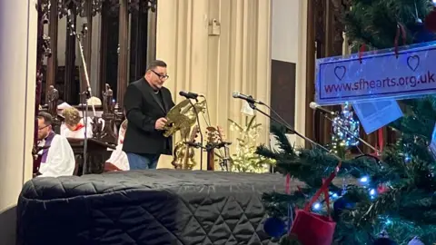 Jon Wright/BBC Wayne Bavin standing behind a gold bird-shaped lectern in a church. There is a Christmas tree in the foreground and the choir is visible behind him