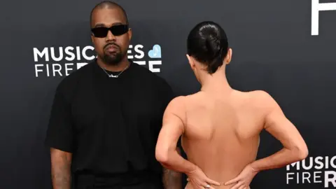 Getty Images Kanye West wearing black opposite his wife wearing very little