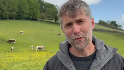 Lee is standing in a field and there are grazing cows in the background he is wearing a grey hooded top and has dark hair and a beard flecked with grey. 