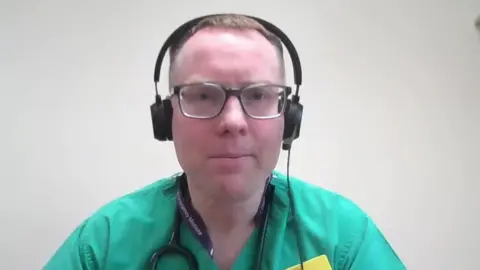 A screenshot of Dr Gareth Hampton from an online interview. He is a younger manm with short, light-coloured hair. He is wearing a black headset with a microphone. He is also wearing green scrubs with a stethoscope around his neck.