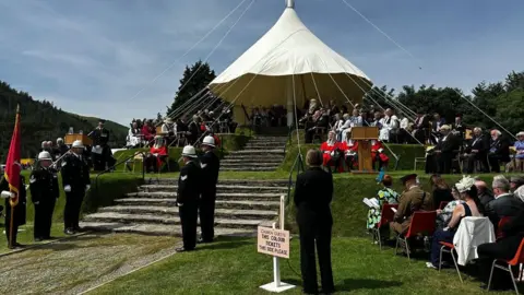 Tynwald Hill, which has a white canopy over the the top rungs. People are seat on each of the tiers and around the bottom.