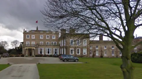 A large building with windows and a flag flying from the roof. It is set back from a driveway and surrounded by grass. 