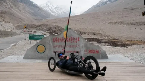 Ben Large A handcyclist lies in a handcycle in front of a stone marker near Everest Base Camp, surrounded by mountainous terrain.