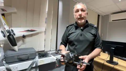 A police officer with grey hair and a beard and wearing a black uniform holds a small black drone. There is a fixed wing drone in the background.