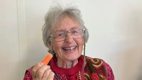 A grey haired older lady with glasses, wearing a colourful cardigan and holding a carrot 