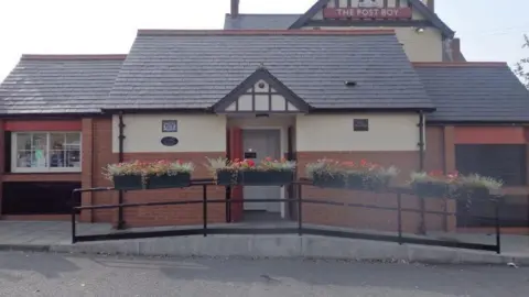 The public toilet is a red-bricked building with a tiled roof and a cream concrete exterior décor. A red door in the middle opens up onto the toilets. There are planters on top of the black metal fence out front.