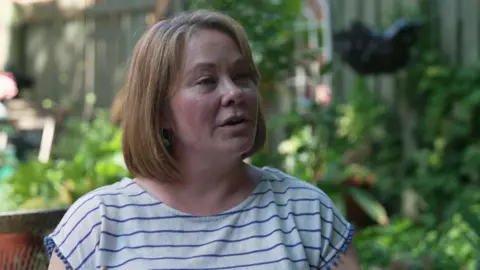 Dr Megan Klaar, a woman with shoulder-length fair hair and a striped t-shirt, sits in a garden. Blurred plants can be seen behind her.