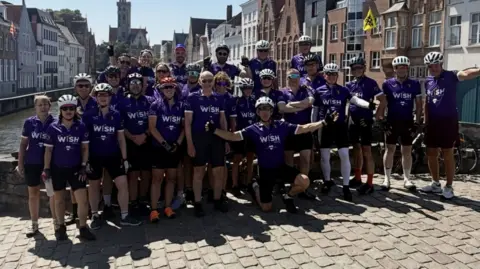 Contributed A group of cyclists stand on a bridge in the middle of a European town. They wear matching purple cycling jerseys with black cycling shorts. They are smiling and some have their arms raised out in celebration. Some have helmets on and sunglasses.