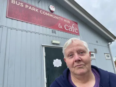 Older woman in a purple and blue top in front of a building with thesign Bus Park Community Hub and Cafe