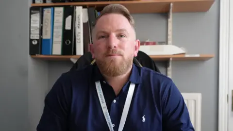 Qays Najm/BBC Ricky Bollden slightly smiles at the camera as he sits in an office. He has light hair with a light beard. He wears a dark blue shirt with a white lanyard around his neck. 