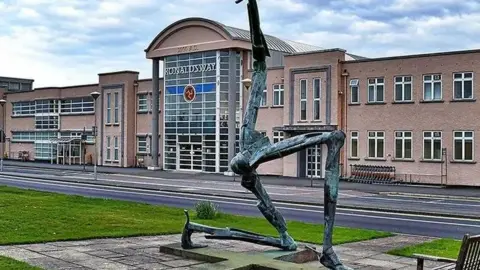 The exterior of the Isle of Man Airport. A three legs of man sculpture can be seen in front of the peach-coloured airport building, with a sign that reads Ronaldsway.