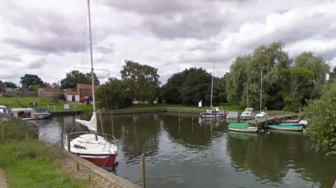 Boats in a marina at Rockland St Mary. Trees line much of the marina and a brick building is in the background.