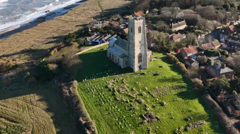 A church viewed from the air, showing its graveyard, fields and the sea beyond, to left, and housing to the right. 