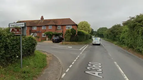 A Google Maps screenshot of a road. Is it a T junction, with a row of two or three red brick houses on the corner. It is in a rural location, with tall hedges and trees along the road. 
