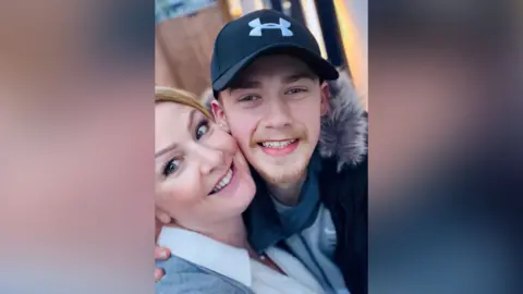 Sue Gray Sue Gray and Matthew Gray. Both are smiling to camera. Sue is wearing a white shirt and grey cardigan. Matthew is wearing a cap and black puffer jacket.