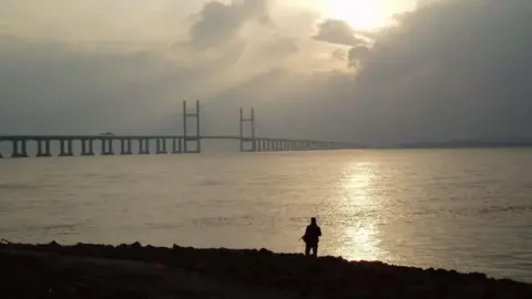 Silhouette of person stood on the bank of the River Severn in front of one of the Severn Bridges as the sun is coming out from behind a cloud.