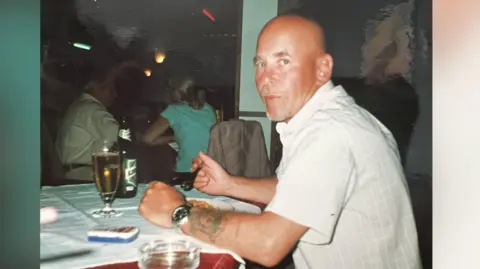 Diane Adams A man is sitting in a restaurant eating a meal with a beer in front of him. He appears to be on holiday and has a sun tan. He is wearing a short sleeved shirt. He has a bald head and appears to be in his mid-50s.