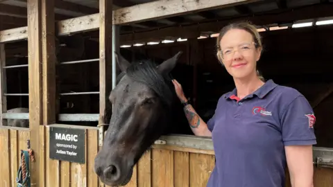 Faye Bedford, who has blonde hair tied back, is photographed wearing a purple polo-neck shirt. She smiles for the camera while stroking a dark brown pony resting its head on stable doors.