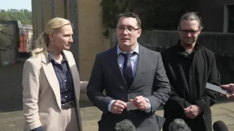 Christopher Trybus wearing a dark grey suit and navy tie, standing outside court delivering a statement to the press, with a folded piece of paper in his hands. A woman wearing a pinstripe shirt and light-coloured suit stands beside him, holding onto his arm.