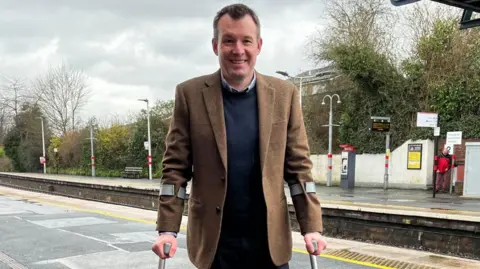 Mr Anderson pictured smiling and looking into the camera. He's wearing a brown tweed blazer, and a navy jumper and trousers. He's also using crutches, with the grey plastic handles just visible. Behind him is Ludlow station platforms, separated by the tracks. The sky is grey and overcast. 