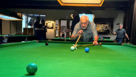 A man with white hair and a check shirt. He is leant over the snooker table poised to make a shot on a green ball.