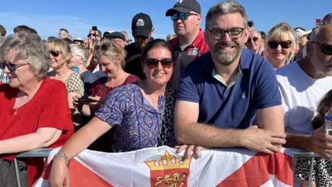 Lucy and Nick Le Boutillier are standing at the front of a crowd at the opening ceremony, leaning on a barrier with a Jersey flag.