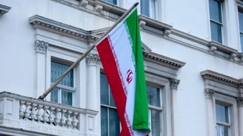 A close-up shot of the upper floors of the Iranian Embassy focusing on an Iranian flag hanging from a pole that sticks out from a balcony