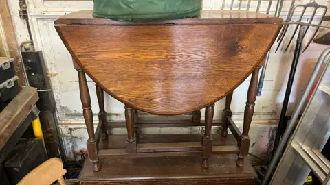 John Devine An oak drop leaf table sits in a garage with another one below it, the wood is a dark brown colour, a step ladder and chairs are also visible.