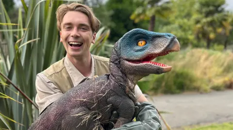 A man, wearing a off white top and light green vest, smiling at the camera holding a velociraptor. The velociraptor is on it's side, it has purple scales on it's body and blue scales on its head and has it's mouth open and a big orange eye. Behind them is green bushes and trees and a concrete path, the background is blurred. 