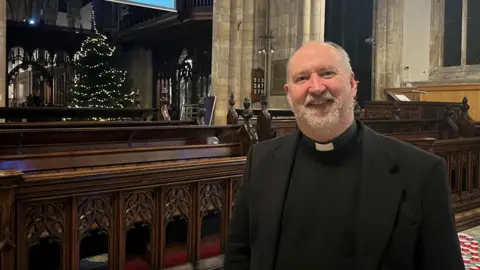 A vicar, wearing his black religious dress and clerical collar, in front of a Christmas tree and church pews. He was short greying hair and beard and is smiling.