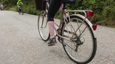 A woman cycling a beige bike on a rural cycle trail. The image is cut off at the woman's waist so all that can be seen is her legs on the pedals. There are more cyclists in the background.