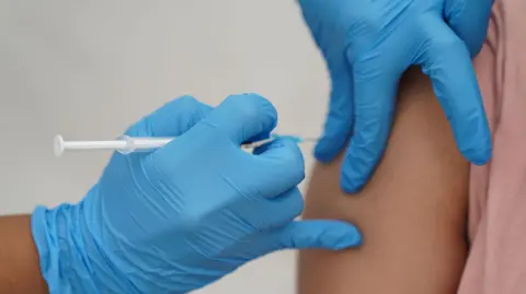 A nurse wearing blue medical gloves injects a vaccination into a patient's upper arm.