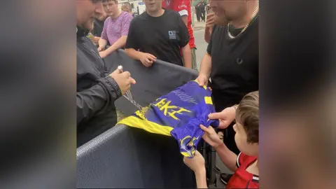 Family photograph Darts star Luke Littler signs a Luke Littler purple and yellow darts top being held by 10-year-old Harry with short brown hair who is wearing a red football shirt with his dad with a brown beard wearing a black t-shirt to the left. They are standing outside Blackpool's Winter Gardens.