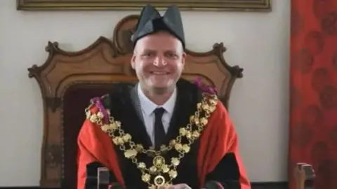 Aigars Balsevics in mayoral attire, a black hat, a gold chain and red robe, with a suit underneath. He is sitting in an ornate wooden chair.