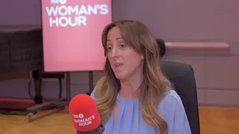 Natalie Cassidy has long wavy brown hair. She is wearing a light purple blouse.