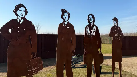 Four rusted steel figures are fixed to the grass and look like steel versions of  cardboard cut-out figures. Each shows a woman in uniform, including pilots and an army sergeant.