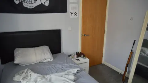 Met Police Inside Coleman's bedroom, which has an unmade bed and a white bedside table next to it. A brown air rifle is leaning against the pale blue wall next to a mirror. A black flag with a skull and crossbones is pinned to the wall above his bed.