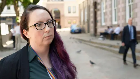 Deputy Doublet has glasses and a green shirt. She is stood in Jersey's Royal Square. 