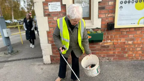 Margaret Walton, die kort wit haar heeft en een donkergroene jas draagt ​​met een hoogvisjasje, heeft een afvalverzamelaar en een plastic emmer vast