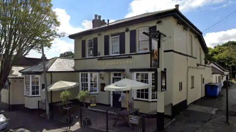 Google The exterior of The Three Colts pub, which is a two-storey, cream coloured building. It has two benches outside, both with umbrellas and enclosed within a black, metal perimeter fence. The building has a door and two windows on the bottom floor, with three windows on the first floor. It is situated on a residential street.