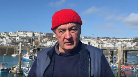 Richard Keen looking at the camera at the Fish Quay with small fishing boats behind him. He is wearing a navy jumper, a blue gillet and a red woolly hat.