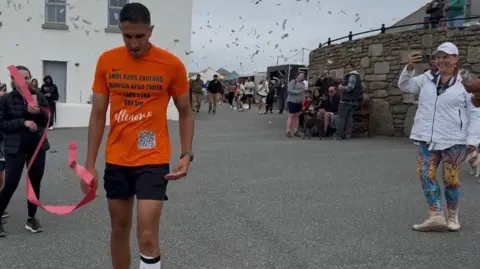 Maisie Randhawa Amol Randhawa crossing the finish line in Land's End, Cornwall. Amol is wearing black shorts and an orange t-shirt with Ellenor emblazoned on the front. A red ribbon and confetti can be seen in the picture. 