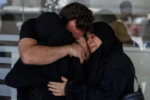 Reuters A man and two women terfully embrace each other outside a hospital in Tyre. The man is wearing a black t-shirt and silver bracelet with his face buried into the shoulder of a woman wearing a black hijab. Both are embraced by another crying woman also wearing a black hijab.