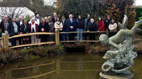 Crawley Borough Council The metal statue of a boy jumping over the top of a fish sits in the middle of a park pond. A large group of people, some wearing chains of office, are lined up along a bridge over the water feature, looking straight at the camera.
