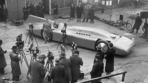 Getty A man in a suit leans against a large space-age looking racing car surrounded by photographers.