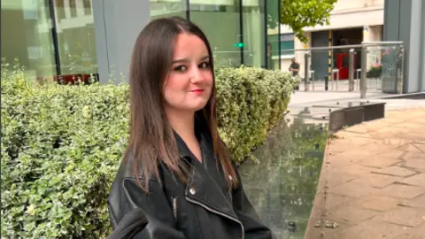 Rachael Williams Lilly is sat on a wall in a black leather jacket smiling at the camera. She hs a green bush or hedge behind her and she is sat on a low, silver-coloured border to the greenery.
