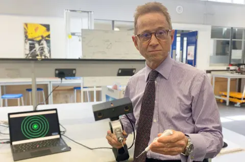 Royal United Hospitals Bath NHS Foundation Trust Professor Grey Giddins, Consultant Orthopaedic and Hand surgeon, holding a new medical device called a 'drill guidance system.'