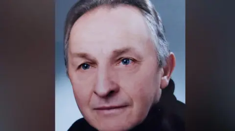 Headshot of a man with a black polo neck on. He has grey swept back hair and blue eyes. 